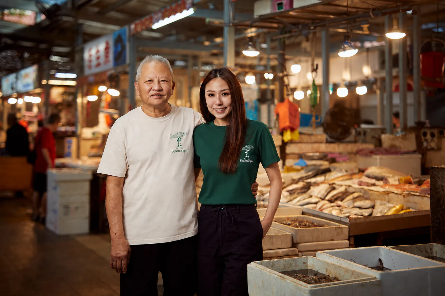Two people in Seamonger shirts at a Singapore wet market, the day's fish laid out on ice beside them.